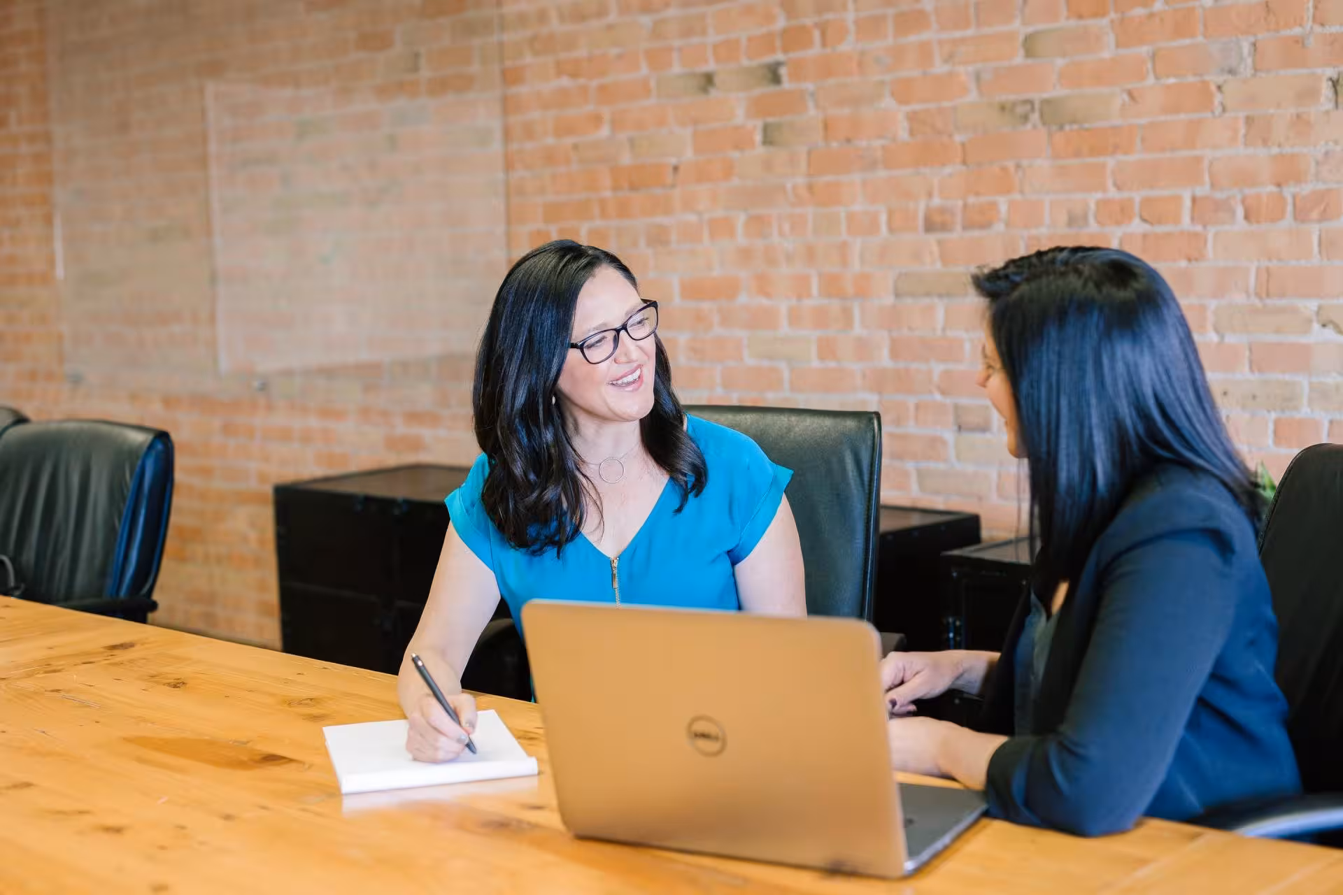 two women speaking to each other taking notes on paper and a laptop